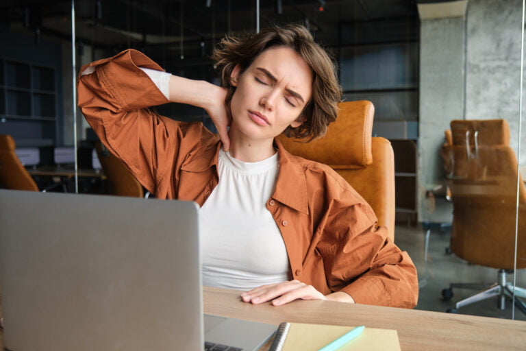 Image of woman with tired face, sits with laptop in office, feels tension in neck, pain in muscles after working all day at workplace